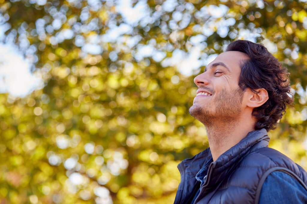 a man wearing a jacket smiles and looks up while standing outside near trees during fall while feeling relief after learning about the stages of addiction recovery