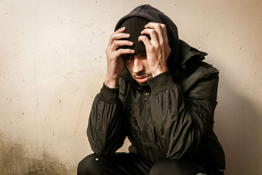 Distraught young man sitting against a wall with his head in his hands hoping someone recognizes the signs of heroin addiction in him before it is too late